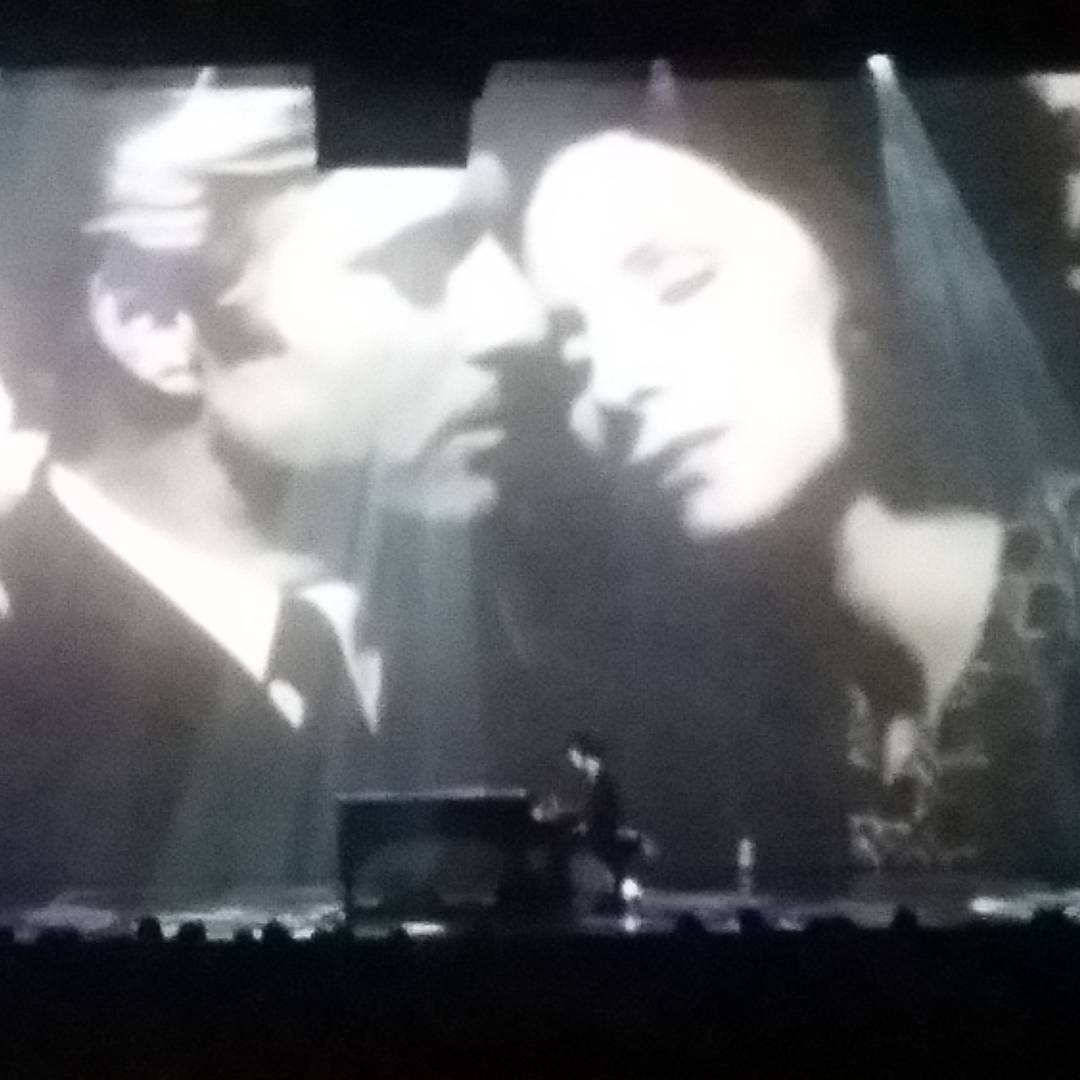 Prince onstage during his final concert, with an image projected behind him of Redford and Streisand from The Way We Were