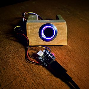 A round fingerprint sensor set into a small wooden block, its ring lit purple, wired to a thumb-sized microcontroller on a workbench.